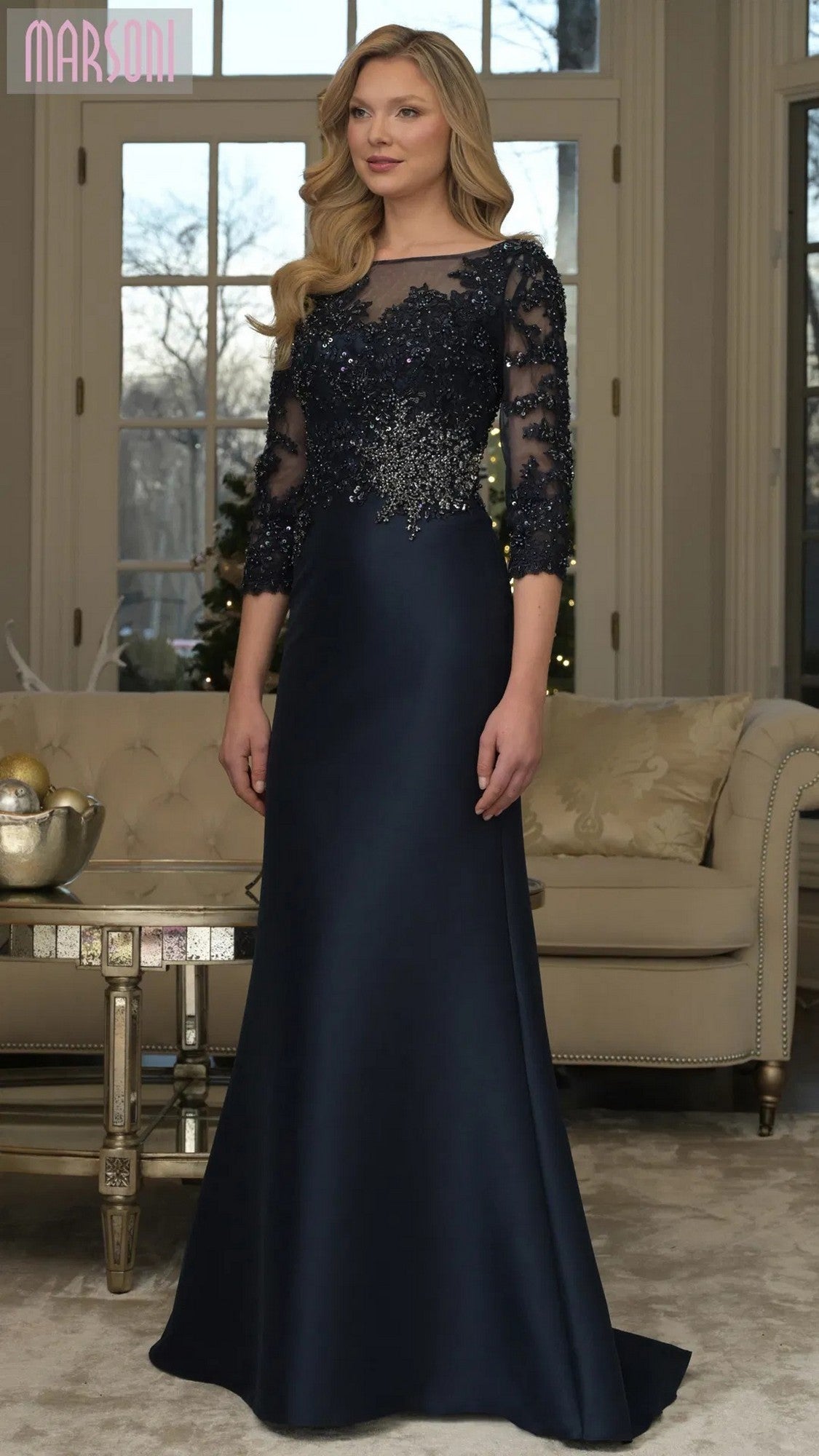 A woman wearing the Marsoni MV1357 long formal dress in navy, featuring a lace-embroidered bodice and sheer three-quarter sleeves, stands in a living room with beige furniture.